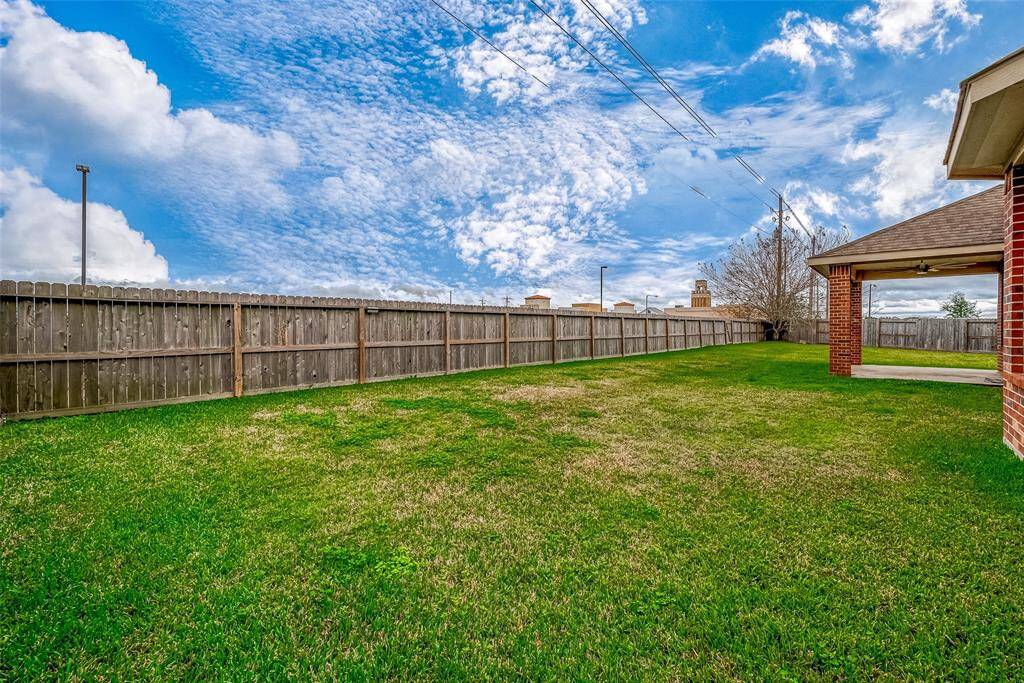 This photo showcases a spacious, well-maintained backyard with a lush green lawn, bordered by a wooden privacy fence. The area is open and sunny, with a partial view of the house's red brick exterior and covered patio, ideal for outdoor activities and relaxation.