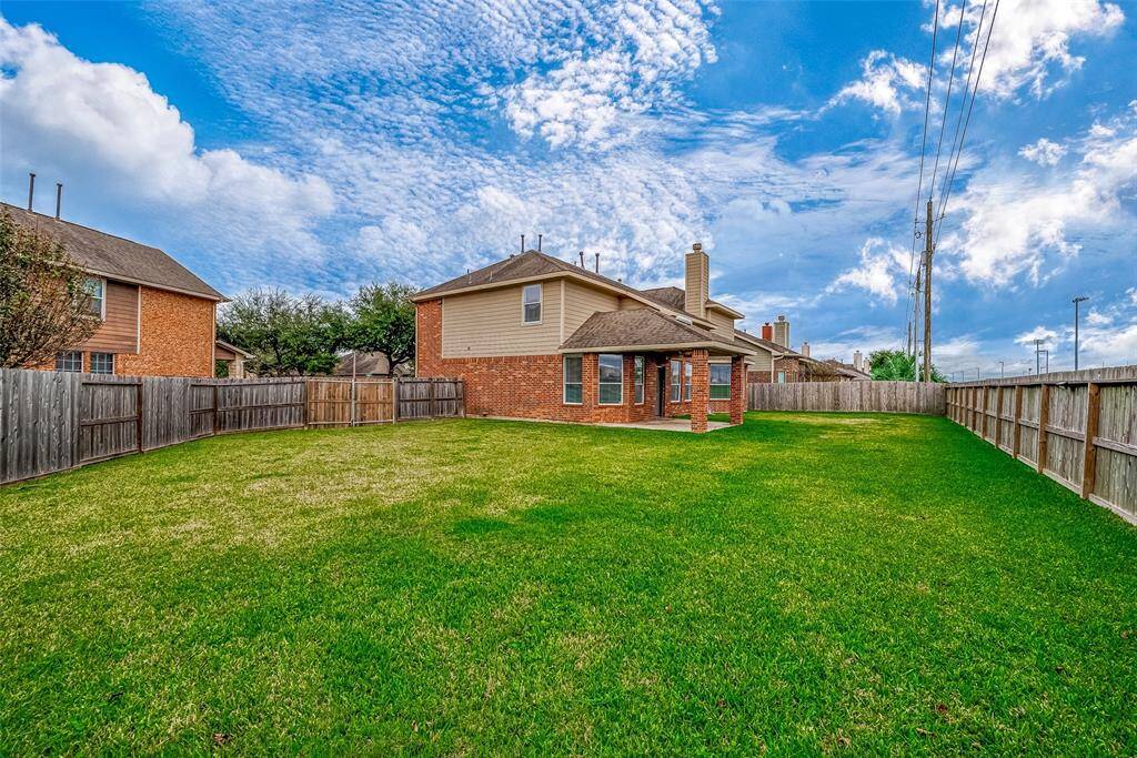 This photo features a spacious backyard with a well-maintained lawn, enclosed by wooden fencing. The two-story brick house has a covered patio, ideal for outdoor activities. The sky is clear, adding a bright and inviting atmosphere.