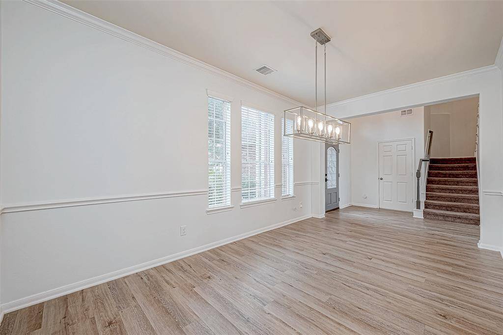 Bright dining area with wood flooring, large windows, and a modern chandelier. It opens to an entryway with stairs and a doorway, creating a welcoming atmosphere.