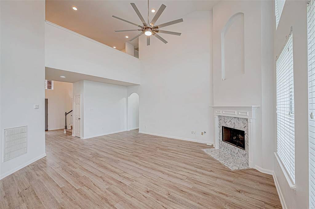This spacious living area features high ceilings, a modern ceiling fan, and abundant natural light. The room includes a stylish fireplace with a herringbone tile design, complemented by elegant wood flooring.