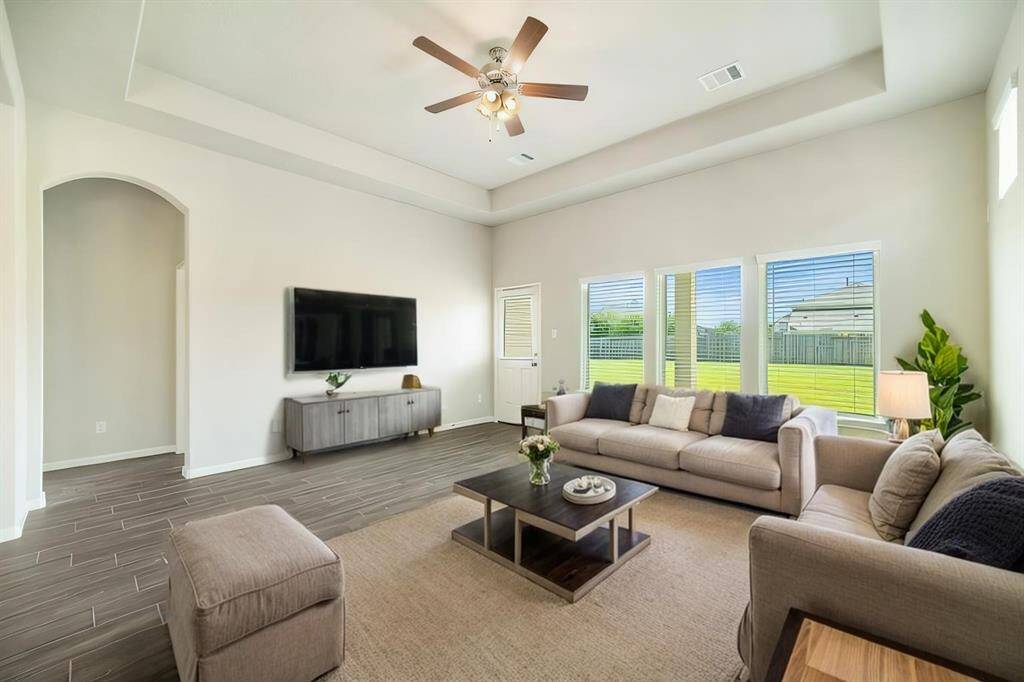 This spacious living room is designed for comfort and elegance, featuring soaring tray ceilings, recessed lighting, and a modern ceiling fan. The large windows flood the space with natural light, offering serene backyard views and a seamless connection to the covered patio. The neutral color palette and wood-look tile flooring create a warm yet sophisticated ambiance, perfect for both relaxing evenings and lively gatherings.
