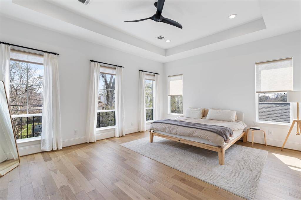 A spacious primary bedroom featuring wide plank wood flooring, tray ceilings, and large windows that fill the space with natural light. Enjoy serene views of the tree-lined neighborhood from this private retreat.