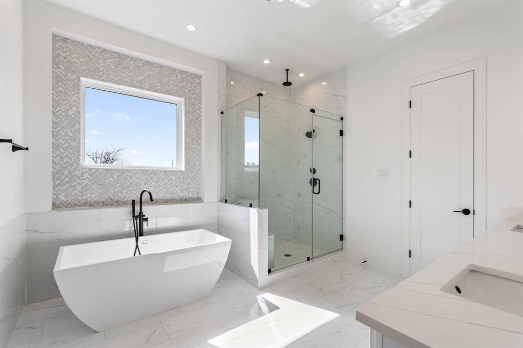 A sophisticated bathroom retreat with a freestanding soaking tub and a glass-enclosed rainfall shower. The textured herringbone tile accent wall and sleek black fixtures create an atmosphere of refined luxury.