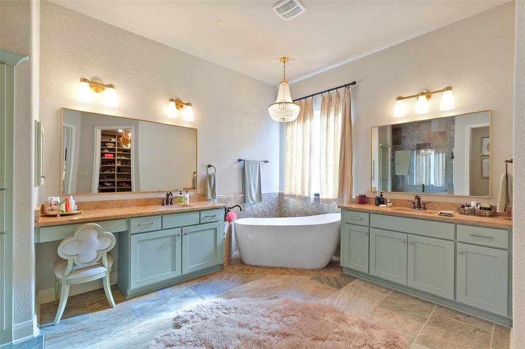 This spacious bathroom features dual vanities with ample storage, a luxurious freestanding tub, and elegant lighting. The soft color palette and large mirrors enhance the airy feel, complemented by a plush rug and natural light. Perfect for relaxation and convenience.