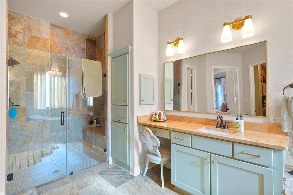 This bathroom features a spacious walk-in shower with natural stone tiles and a glass door. It has a large vanity with a granite countertop, dual sinks, and ample cabinet storage. Warm lighting fixtures and neutral tones create a cozy, elegant atmosphere.