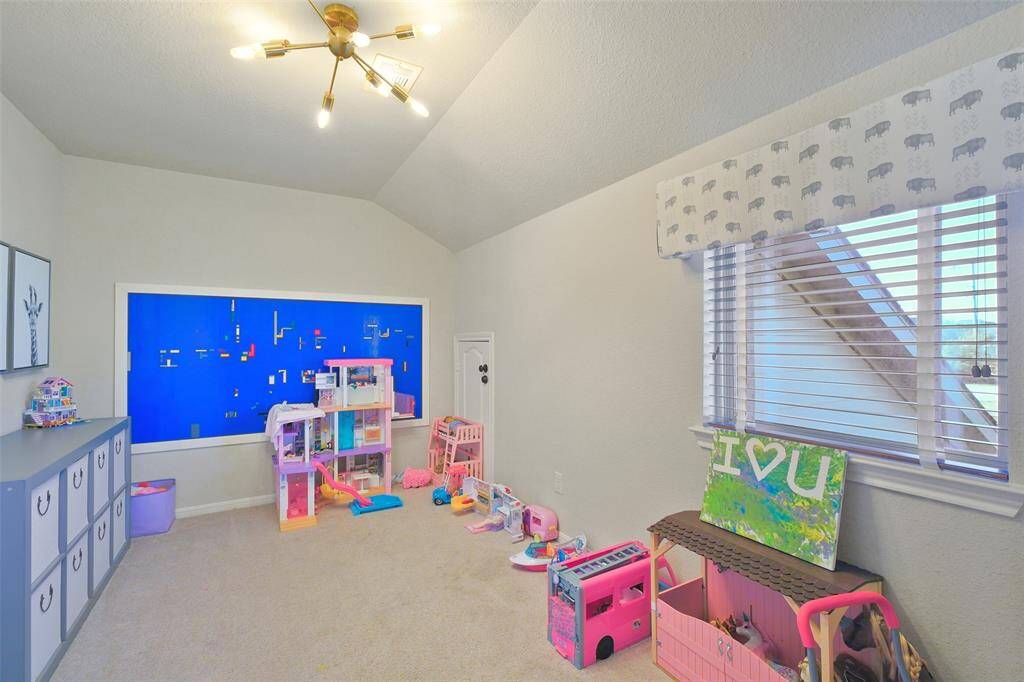 Hidden room behind the bookshelf opens up to a bright playroom with a vaulted ceiling, modern light fixture, and large window with blinds.