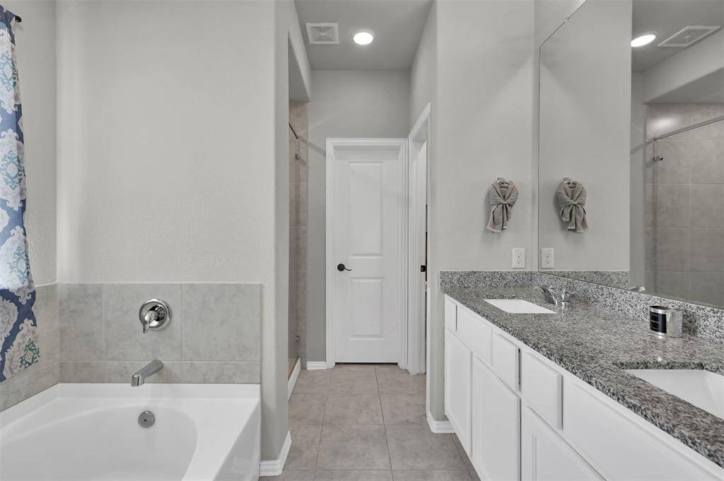 This bathroom features a modern design with a granite countertop double vanity, a spacious bathtub, & a tiled shower