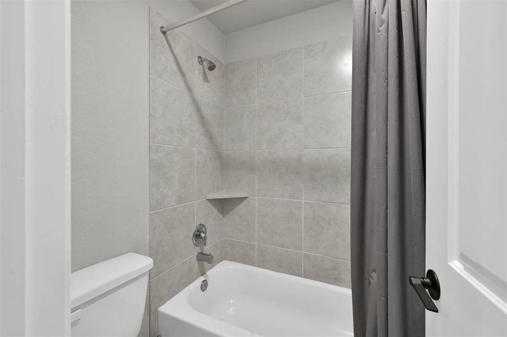 This bathroom features a clean, modern design with light gray tiled walls surrounding a white bathtub & includes a standard toilet, offering a simple and functional setup