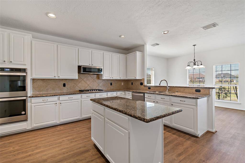 This kitchen features modern stainless steel appliances, ample white cabinetry, and a large island with granite countertops. The open layout and wood flooring provide a warm and inviting space, complemented by natural light from two large windows.