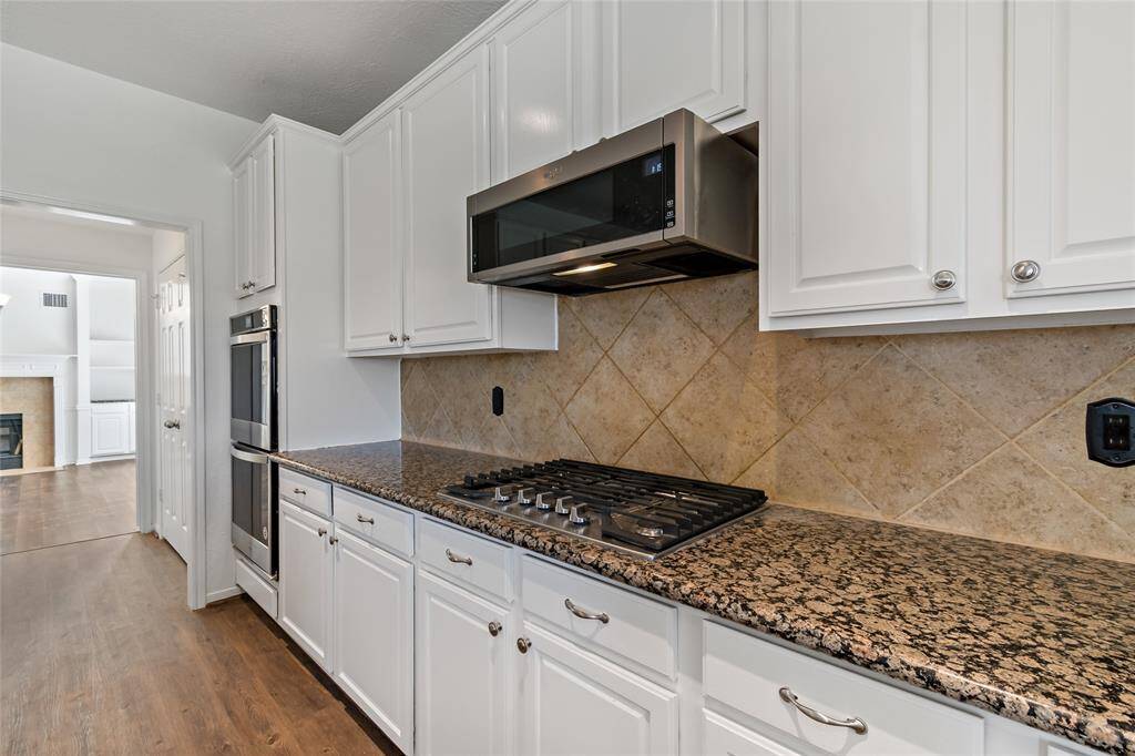 This kitchen features elegant white cabinets, a granite countertop, and a built-in gas stove. The space includes a stainless steel microwave and oven, with a tiled backsplash for a modern touch. The adjacent room with a fireplace adds cozy charm.