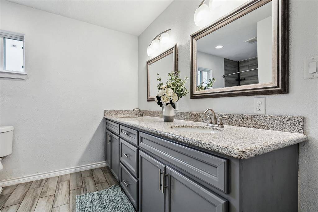 The primary bathroom features a spacious double vanity with granite countertops and stylish gray cabinetry. It includes two large framed mirrors and modern light fixtures, with a neutral color palette that enhances the room's bright and inviting atmosphere.
