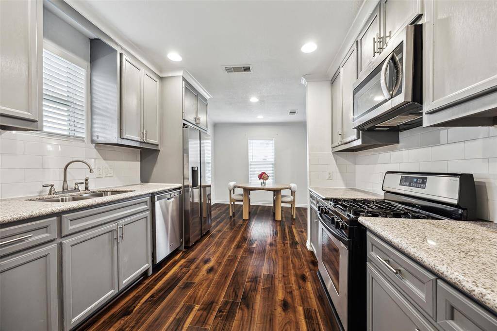 This kitchen features sleek granite countertops, stainless steel appliances, and modern gray cabinetry. The space is well-lit with recessed lighting and has rich hued flooring that adds warmth. A dining area is visible at the end, creating an open and inviting atmosphere.