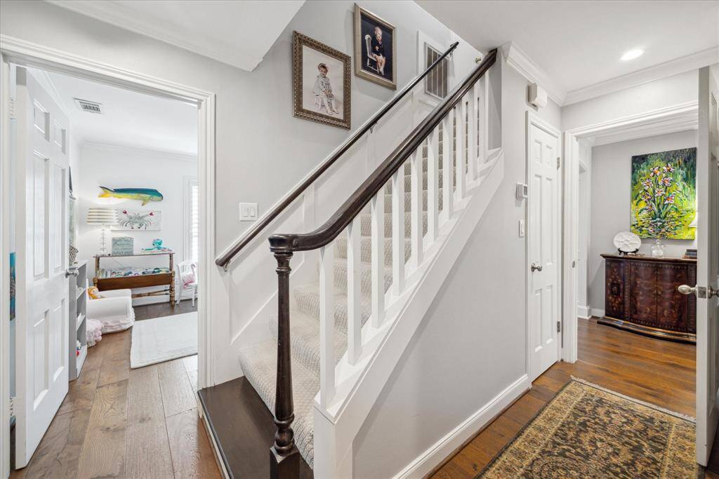 This photo shows a well-lit hallway with a staircase featuring a dark wooden handrail and white balusters. The area has light walls and  hardwood floors. .