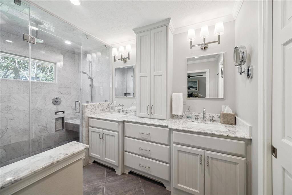 The ensuite primary bathroom features a modern design with a spacious glass-enclosed shower and elegant marble tiles. It has a double vanity with white cabinetry and a large mirror, providing ample storage and a bright, clean aesthetic.