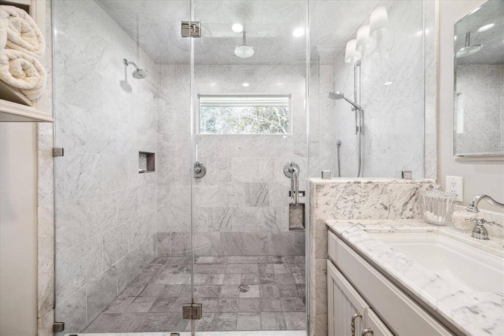 This bathroom features a spacious, luxurious walk-in shower with glass doors and elegant marble tiles throughout. It includes dual showerheads, a built-in shelf, and a sleek vanity with a marble countertop.