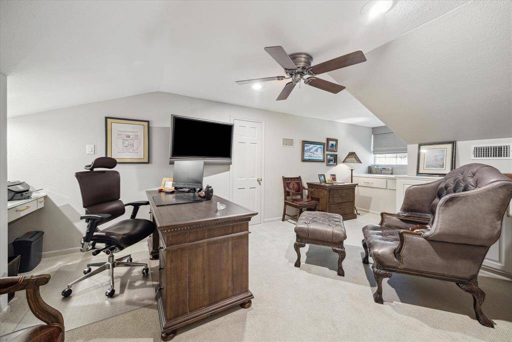 This photo showcases a cozy home office with sloped ceilings, featuring  a ceiling fan, and ample natural light. The space includes a comfortable seating area ideal for a productive and stylish work environment.