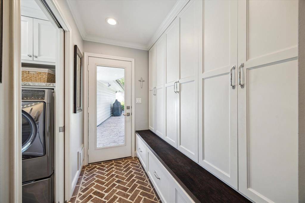 This photo showcases a bright and functional mudroom with ample white cabinetry for storage, and a stylish herringbone tile floor. A door leads to the outdoor patio, and a glimpse of a laundry area is visible, offering convenience and organization.