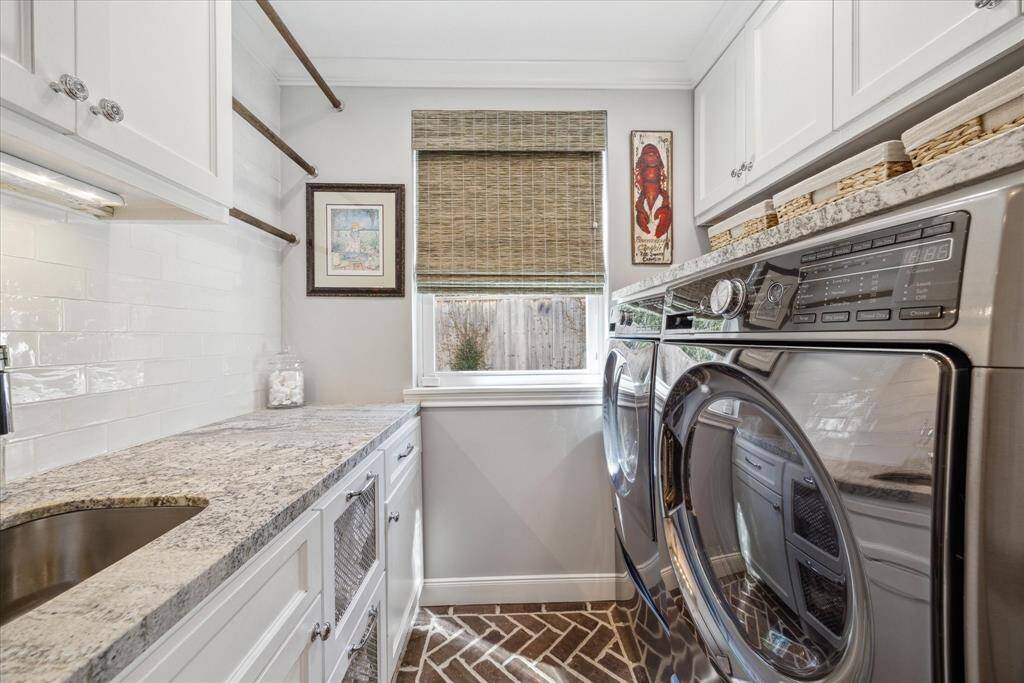 This laundry room features sleek stainless steel appliances, granite countertops, sink,  and ample cabinetry for storage. A window provides natural light, complemented by stylish decor and a textured floor for added warmth.