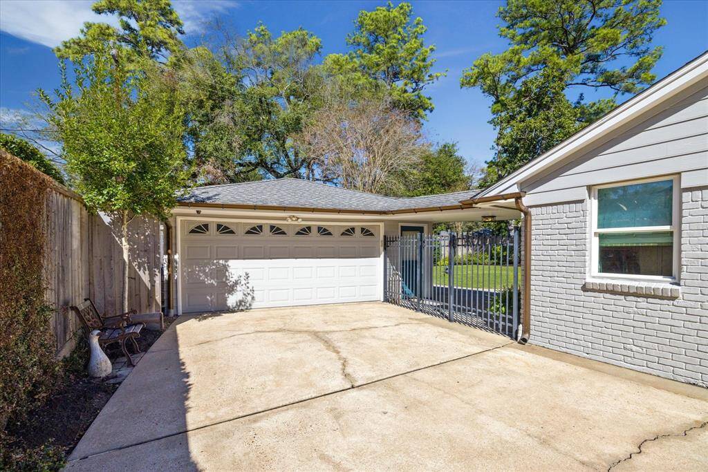 The photo shows a welcoming driveway leading to a double garage with a light gray exterior. There's a small fenced patio area, surrounded by greenery and a wooden fence, providing a cozy and private space. Behind the garage is a shed with window ac and doggie door!