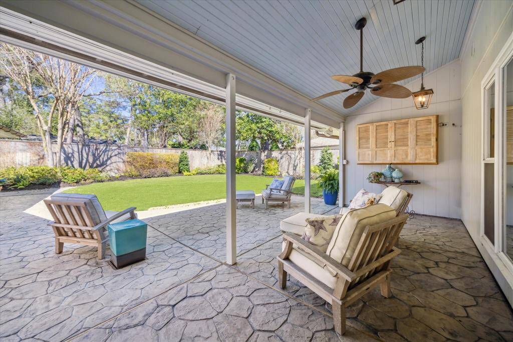 A spacious covered patio with stone flooring and comfortable seating overlooks a lush, well-maintained backyard. The area features a ceiling fan and television cabinet making it ideal for relaxing or entertaining.
