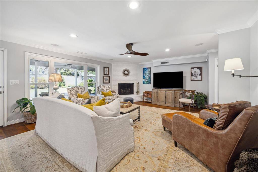 This inviting living room features a cozy fireplace, large windows offering plenty of natural light, comfortable seating, and a ceiling fan. The space is tastefully decorated with neutral tones and accents, providing a comfortable and welcoming atmosphere.