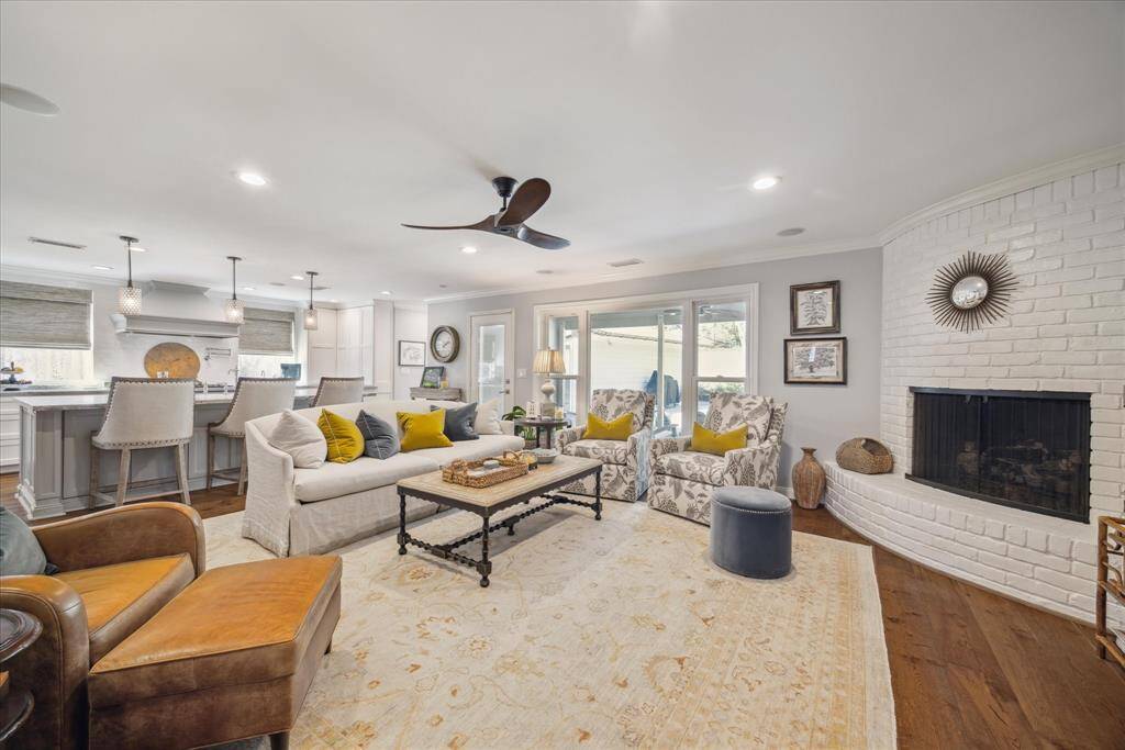 This spacious living area features an open-concept design with a cozy white brick fireplace, hardwood floors, and ample natural light. The room flows seamlessly into a modern kitchen with a large island and bar seating. The decor is stylish and inviting, perfect for entertaining or relaxing.