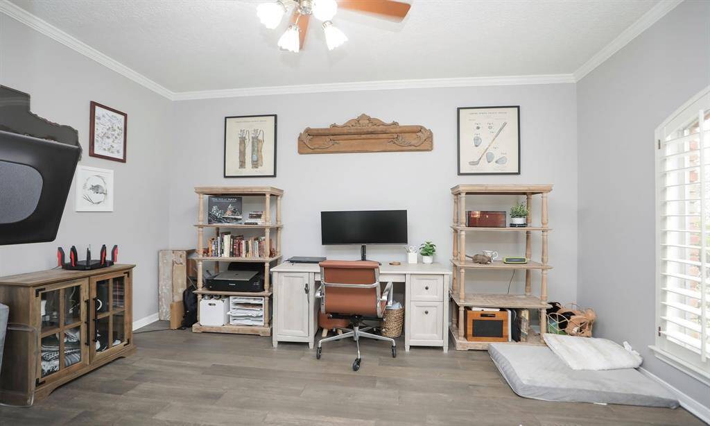 The study features light walls, wood look flooring, and a ceiling fan, creating a comfortable workspace. Large windows with shutters provide natural light.