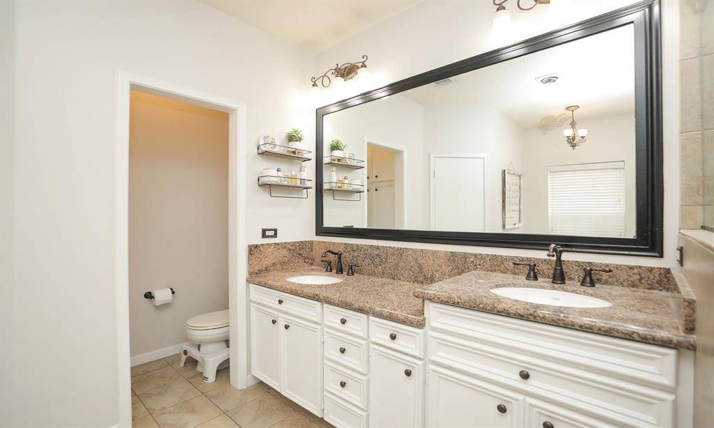 This bathroom features dual sinks with granite countertops, a large framed mirror, and elegant lighting fixtures. It includes a separate toilet area and has stylish wall shelves for storage.