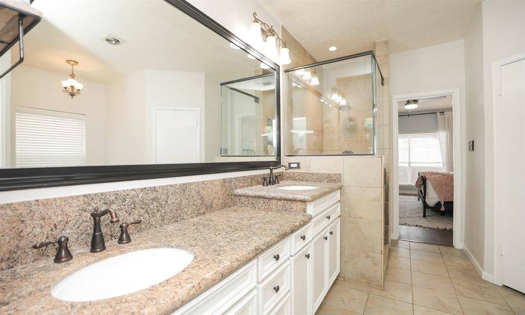 The spacious bathroom with dual sinks, granite countertops, and elegant bronze fixtures. There's a large mirror above the vanity and a glass-enclosed shower. The room leads into the primary bedroom.