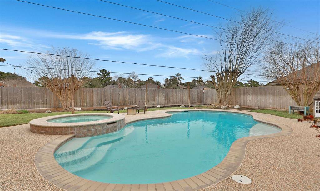 This backyard features a spacious in-ground pool with a connected hot tub, surrounded by a textured patio. The area is enclosed by a wooden privacy fence creating a serene and private outdoor retreat.