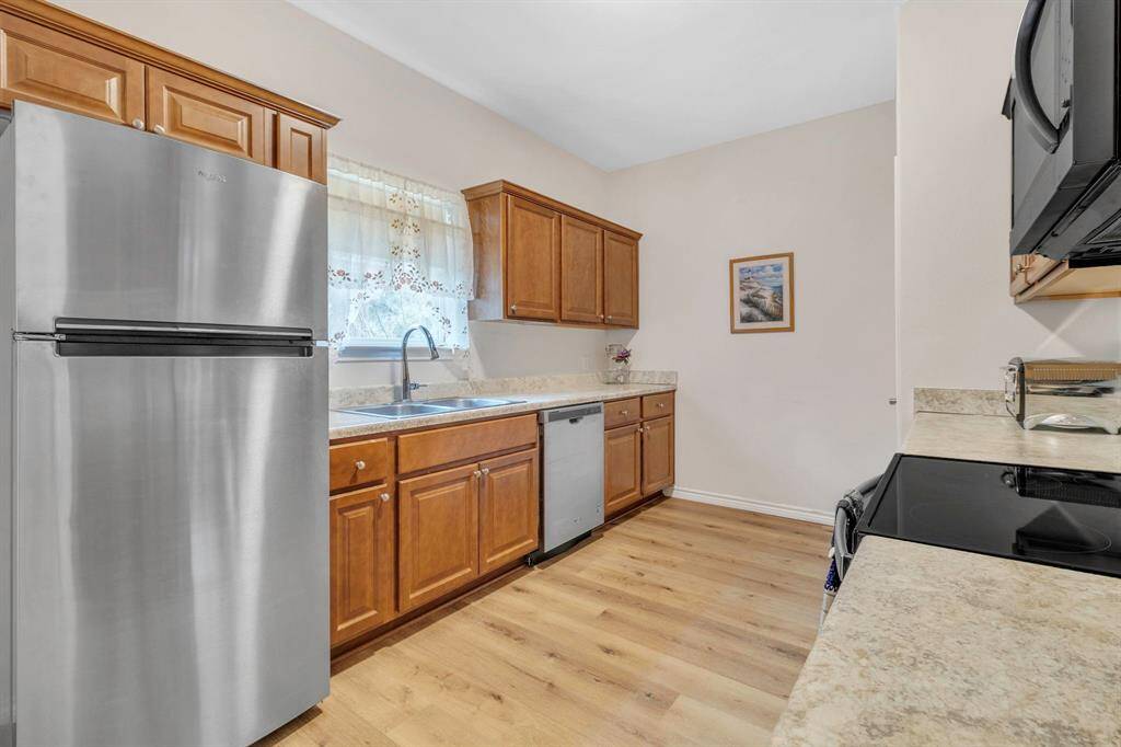 This inviting kitchen boasts stainless steel appliances, warm wooden cabinets, and light countertops. Enjoy the double sink beneath a window with decorative curtains, complemented by wood flooring and a neutral color scheme.