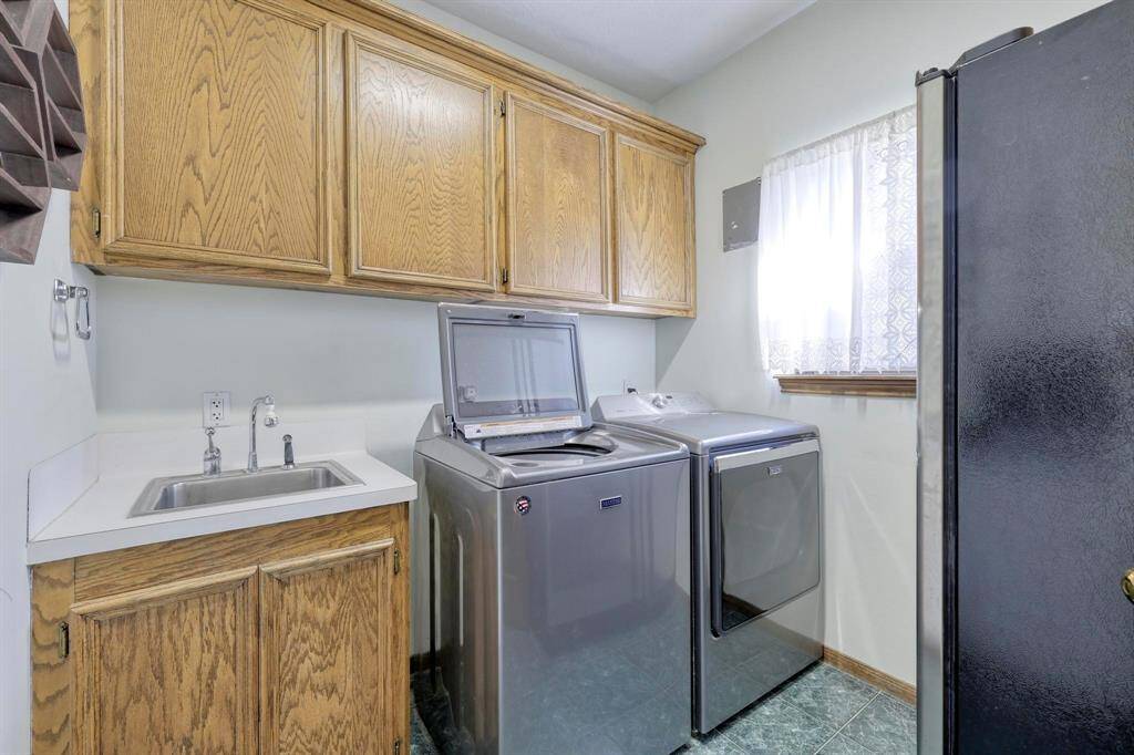 This photo shows a well-organized laundry room featuring modern washer and dryer units, a utility sink with a countertop, and ample wooden cabinet storage. The space is brightened by a window with a sheer curtain, providing natural light.