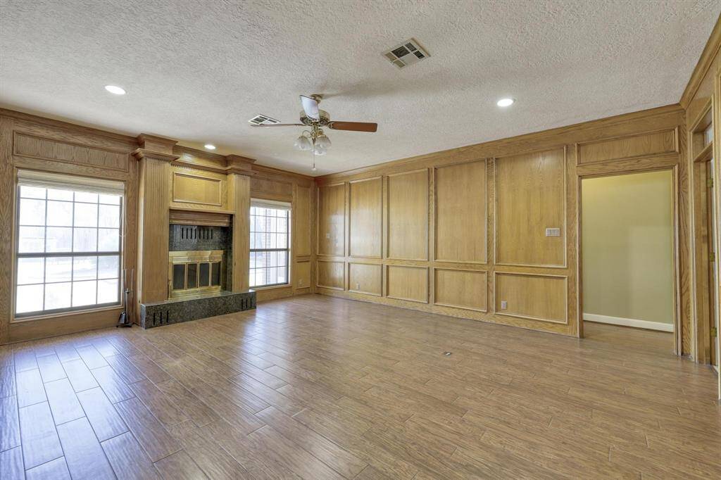 This spacious living room features wood paneled walls, a ceiling fan, and recessed lighting. Large windows provide ample natural light, and a fireplace with a dark stone base adds warmth and character to the space. The room has a cozy yet sophisticated feel.