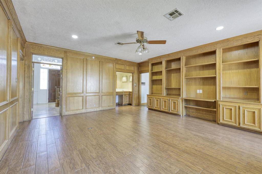 This spacious room features elegant wood paneling, built-in shelves, and a ceiling fan. It has ample natural light and connects to other living areas, offering a versatile space for a home office or library.