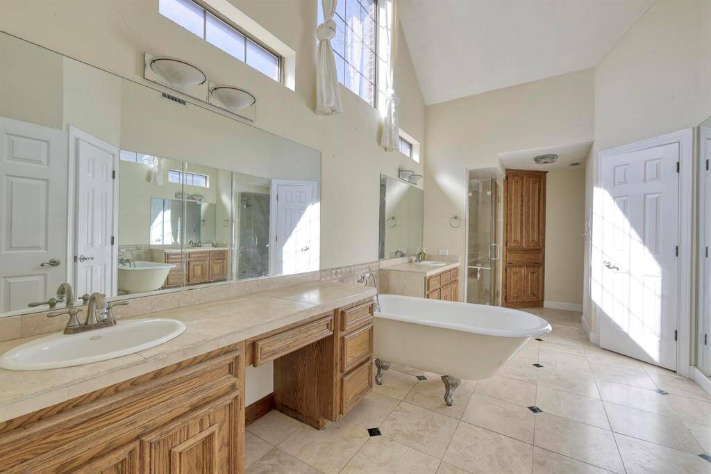 This spacious bathroom features high ceilings and ample natural light from large windows. It includes a classic clawfoot tub, dual sinks with expansive mirrors, and plenty of wooden cabinetry for storage. The neutral tile flooring adds a touch of elegance.