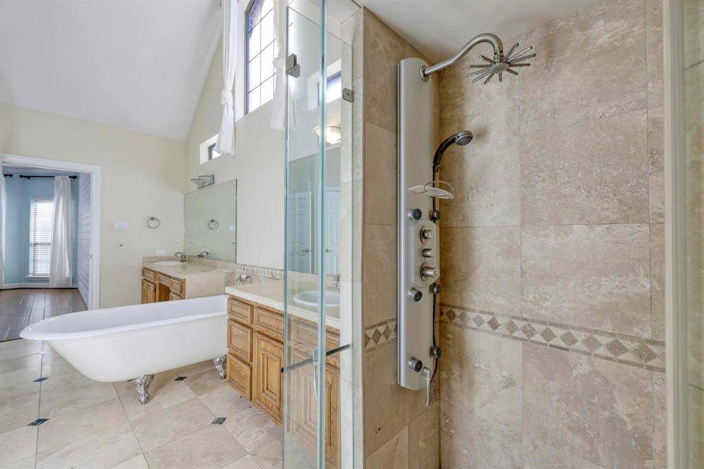 Bright and spacious bathroom featuring a vintage clawfoot tub, dual sinks with wooden cabinetry, and a modern glass-enclosed shower with full-body jets. High ceilings and large windows enhance the airy feel.
