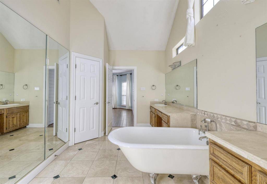 This spacious bathroom features high ceilings, a classic clawfoot tub, dual sinks with ample counter space, and large mirrors. There's plenty of natural light and a direct view into an adjacent room with wood flooring.
