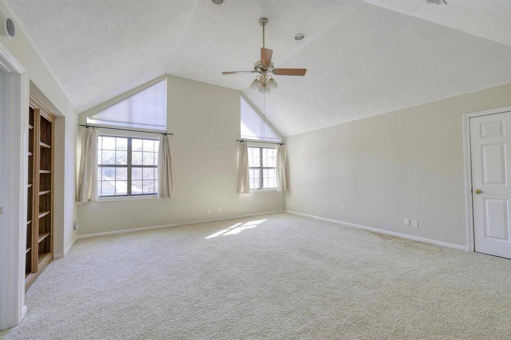 This spacious, carpeted bedroom features a high vaulted ceiling with a fan, large windows for natural light, and built-in shelving for storage.