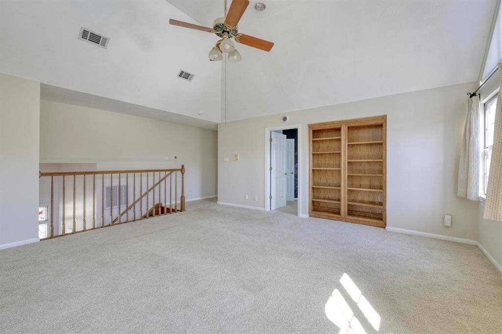 This spacious, carpeted room features a ceiling fan, built-in wooden shelving, and ample natural light from a large window. It includes an open staircase with railing and neutral walls. Ideal for a cozy living area or office space.