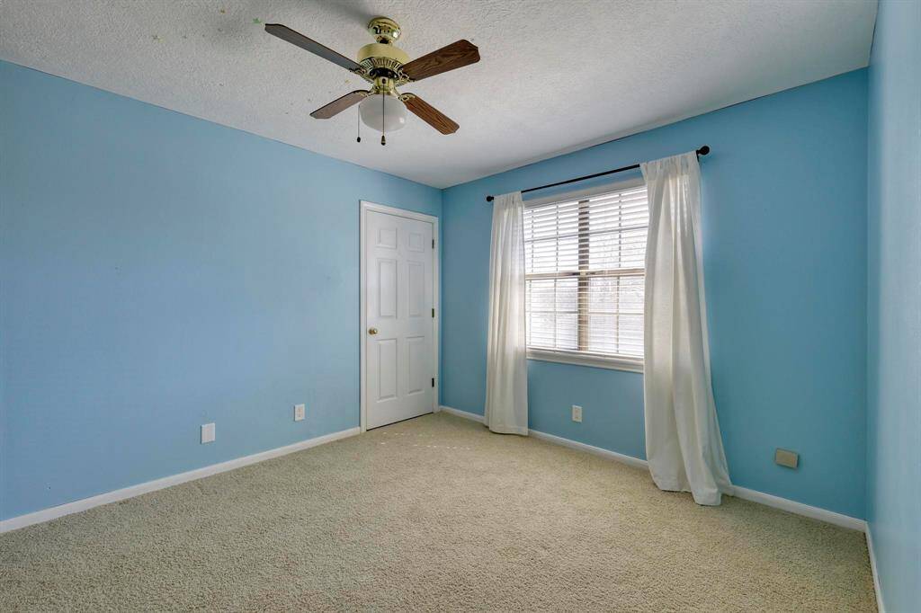 This photo shows a bright, carpeted bedroom with light blue walls, a ceiling fan, and a window with white curtains. A closet with a white door is also visible.