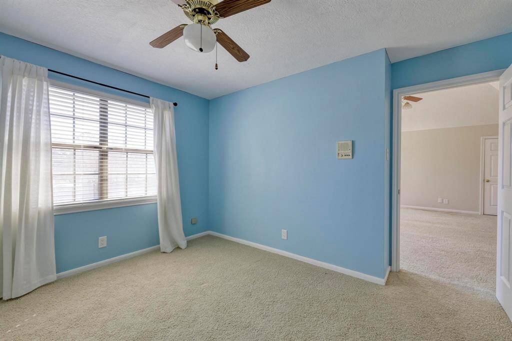 This room features light blue walls, beige carpet, and a window with white curtains, providing a bright and airy feel. A ceiling fan adds comfort, and an open doorway leads to an adjoining space.