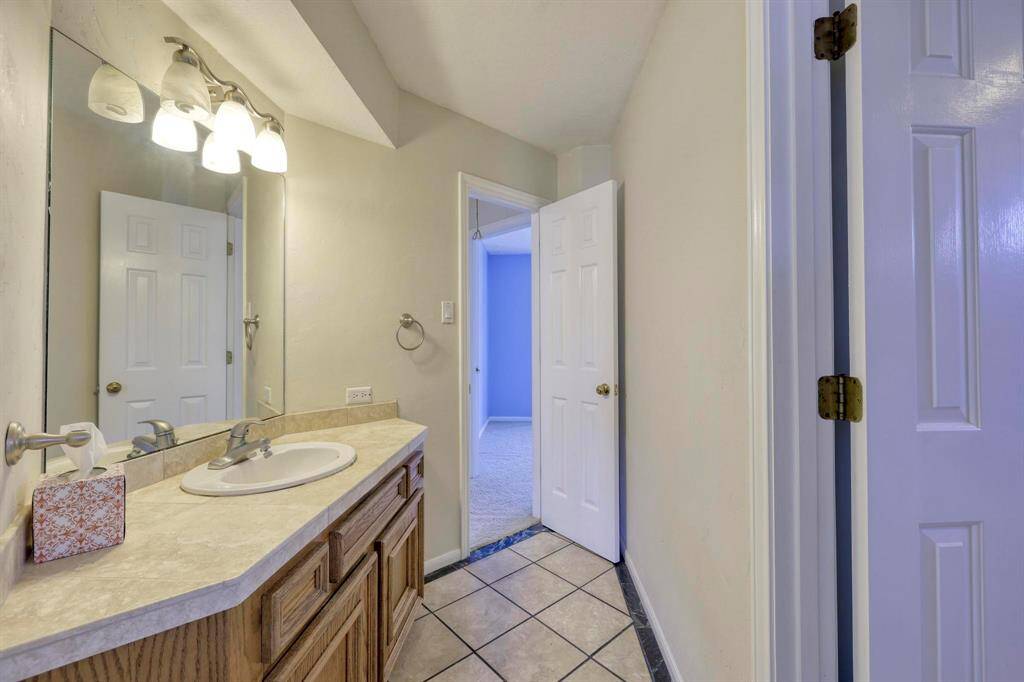 This bathroom features a single vanity with a wide mirror, ample counter space, and wooden cabinetry. It has tile flooring and leads into a carpeted room with blue walls through an open door.