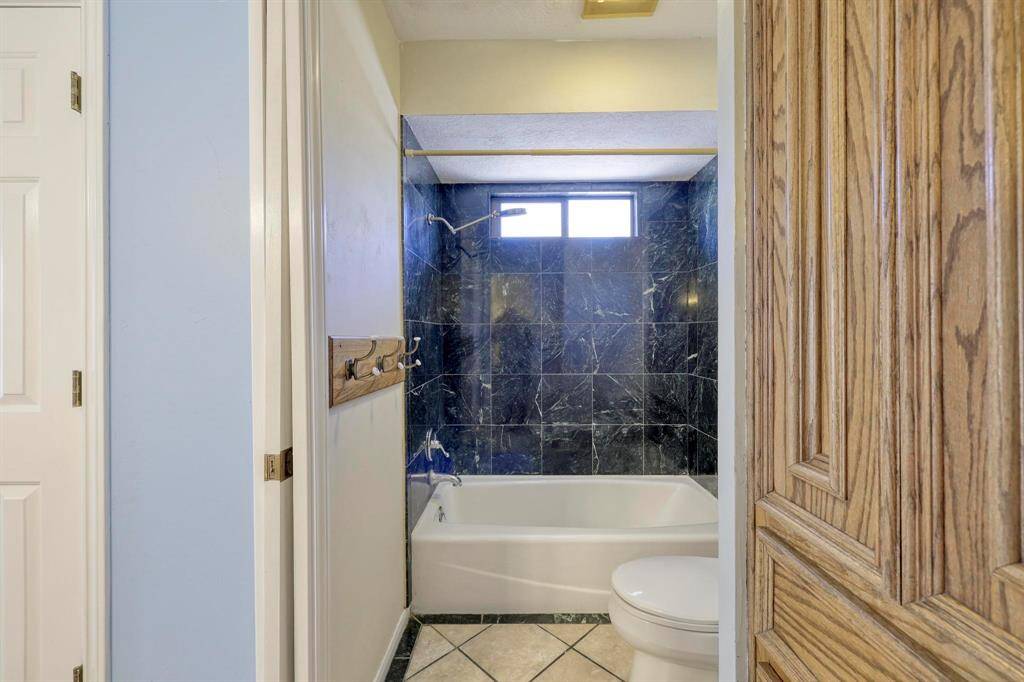 This bathroom features a bathtub with elegant dark marble tiles and a window above. It has light-colored walls, a wooden towel rack, and a wooden cabinet for storage. The floor is tiled with a contrasting pattern.