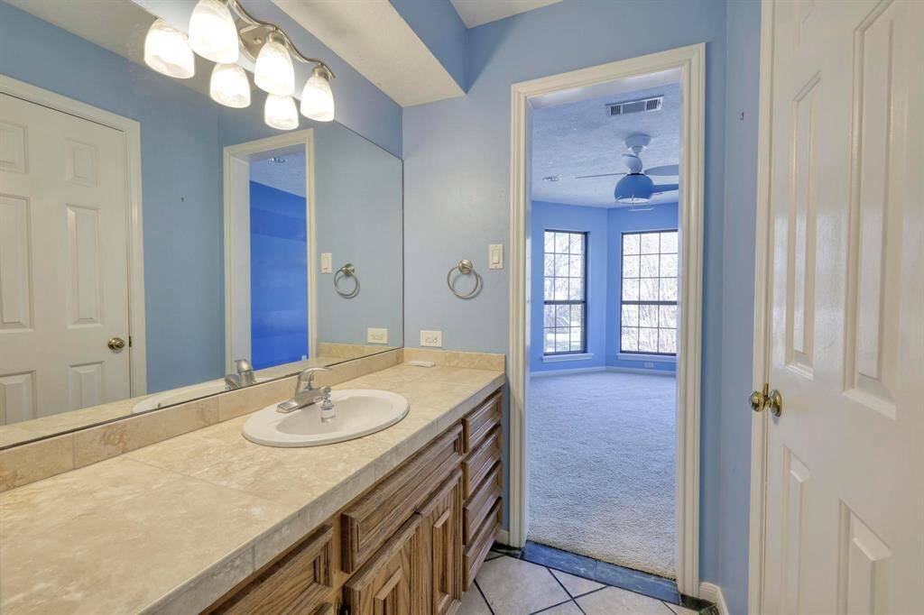 This photo features a bright bathroom with a large mirror, stylish lighting, and a spacious countertop. It opens into a well-lit bedroom with large windows and a ceiling fan, offering a cozy and inviting atmosphere.