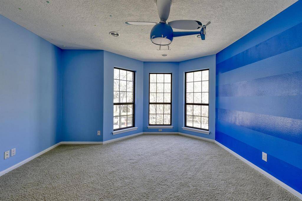 This room features a bright, modern design with light and dark blue walls, a ceiling fan, and large windows offering ample natural light. The carpeted floor adds warmth and comfort.