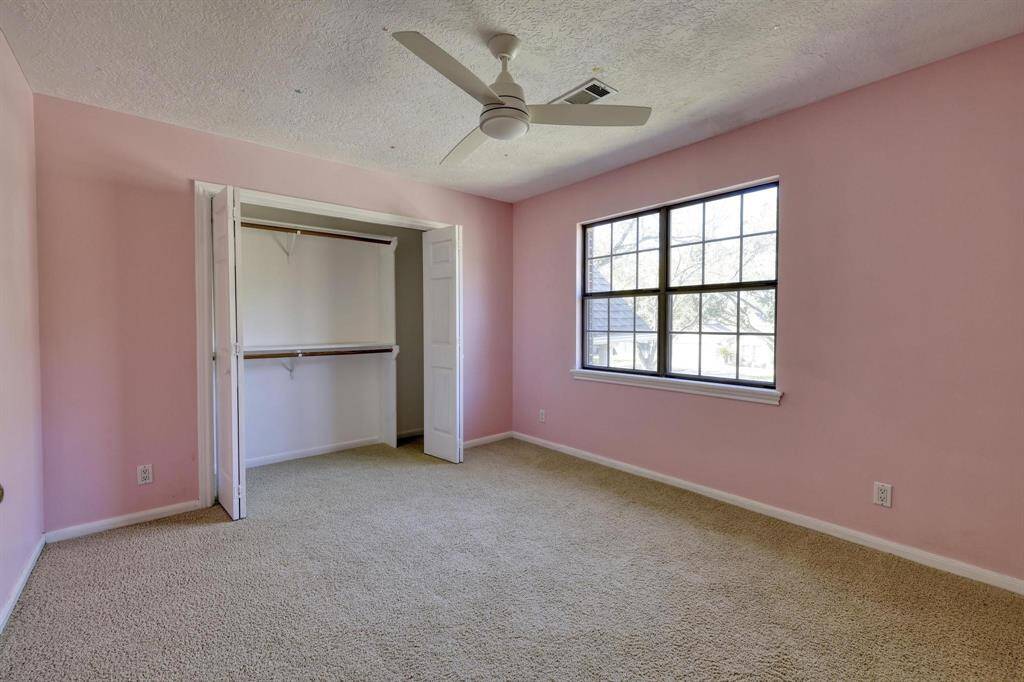 This is a cozy bedroom with soft pink walls and beige carpet. It features a ceiling fan, a window providing natural light, and a spacious closet with sliding doors.