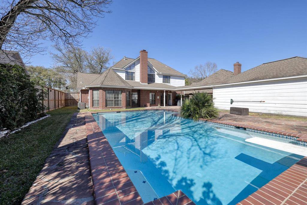 This photo showcases a spacious backyard with a large in-ground pool and a brick patio. The two-story house features a brick exterior and ample windows, providing a bright and welcoming atmosphere. The property is bordered by a wooden fence and includes a detached garage, offering privacy and convenience.