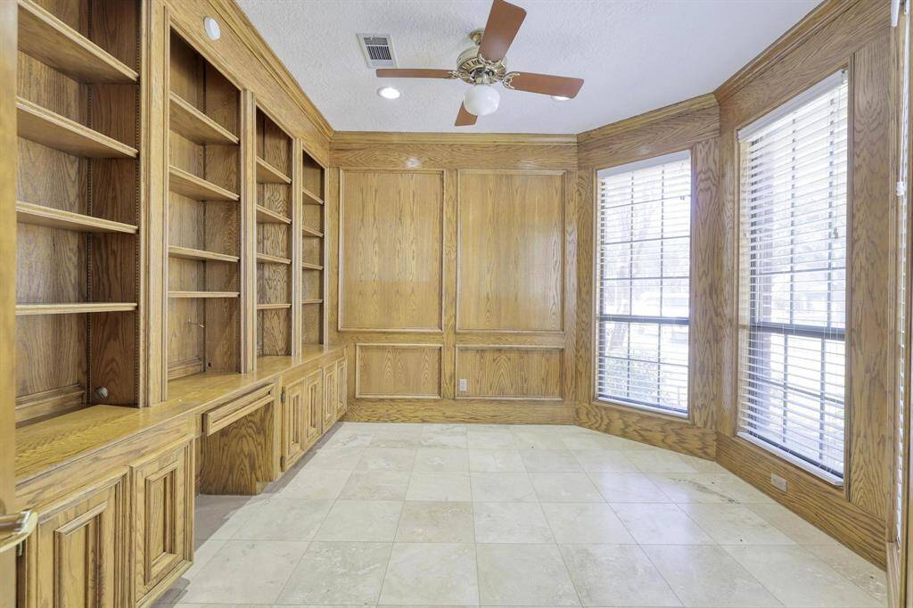 This room features built-in wooden bookshelves and cabinetry, large windows with blinds, a ceiling fan, and tile flooring, creating a bright and functional space ideal for a home office or library.