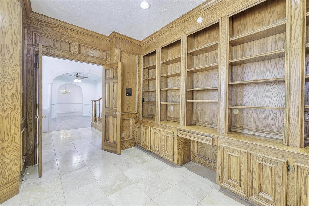 This photo showcases a room with elegant built-in wooden shelving and cabinets, offering ample storage and display space. The flooring is tiled, and an open doorway leads to a bright area with an arched entrance, ceiling fan, and chandelier. Perfect for a home office or study.
