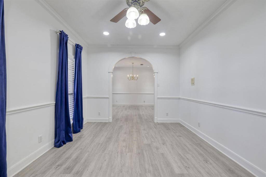 This photo showcases a bright, elongated room with light wood flooring and white walls, accented by deep blue curtains. An archway leads to the dining room, featuring a modern chandelier, adding elegance to the open layout. Ceiling lights and fan provide additional lighting and ventilation.